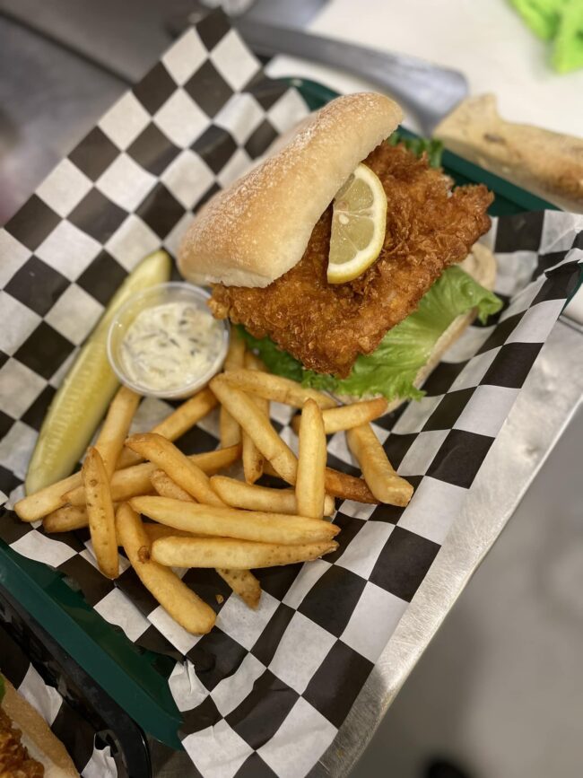 Close-up of a crispy fish sandwich at Fairmount Cafe, featuring a golden, breaded fish fillet nestled in a soft, toasted bun. The sandwich is topped with fresh lettuce, ripe tomato slices, and a dollop of creamy tartar sauce. Served on a rustic wooden table, the sandwich exudes a delicious, hearty appeal.