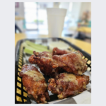 Close-up of a plate of crispy, golden-brown chicken wings at Fairmount Cafe, perfectly seasoned and served with a side of dipping sauce. The wings are garnished with fresh herbs, adding a touch of color and flavor to the dish. The plate sits on a rustic wooden table, highlighting the cafe's warm and inviting atmosphere.