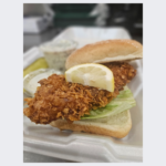 Close-up of a crispy fish sandwich at Fairmount Cafe, featuring a golden, breaded fish fillet nestled in a soft, toasted bun. The sandwich is topped with fresh lettuce, ripe tomato slices, and a dollop of creamy tartar sauce. Served on a rustic wooden table, the sandwich exudes a delicious, hearty appeal.